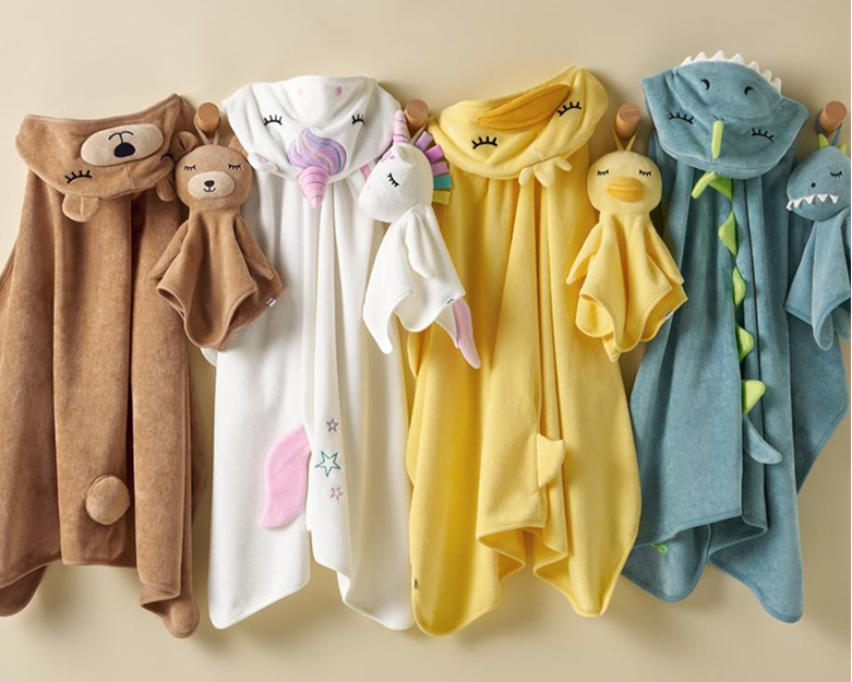 Four plush hooded baby towels in brown, white, yellow, and blue, each with a cute animal face and small plush animal attachment, hanging on a beige wall.