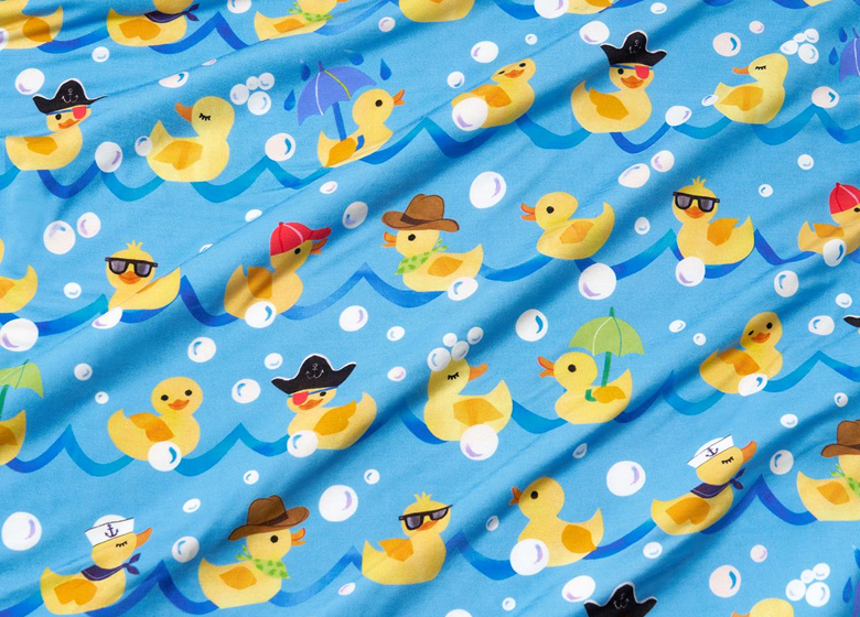 A close-up of a blue fabric with a playful pattern of yellow rubber ducks floating in wavy blue water, decorated with white bubbles and small accessories like hats, sunglasses, umbrellas, and bandanas.