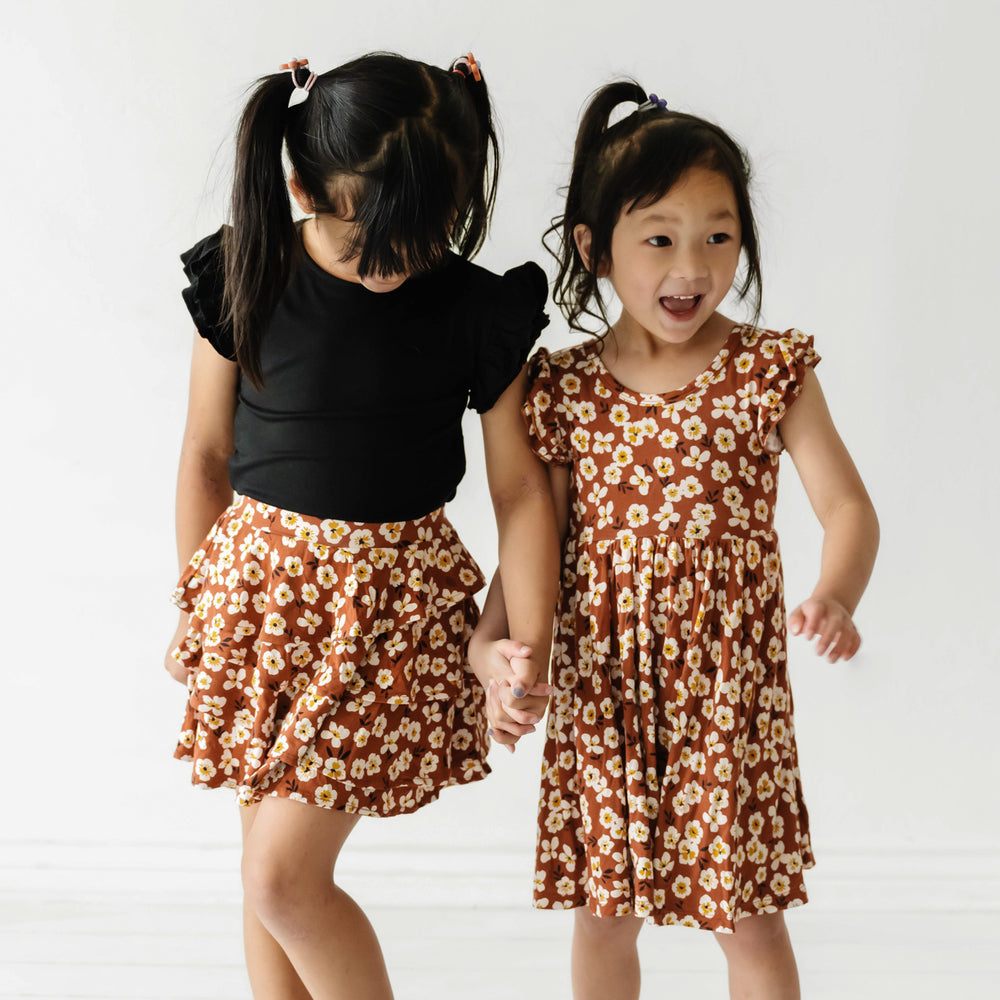 Click to see full screen - Two children holding hands wearing matching Mocha Blossom printed twirl dress and ruffle skirt with black flutter tee