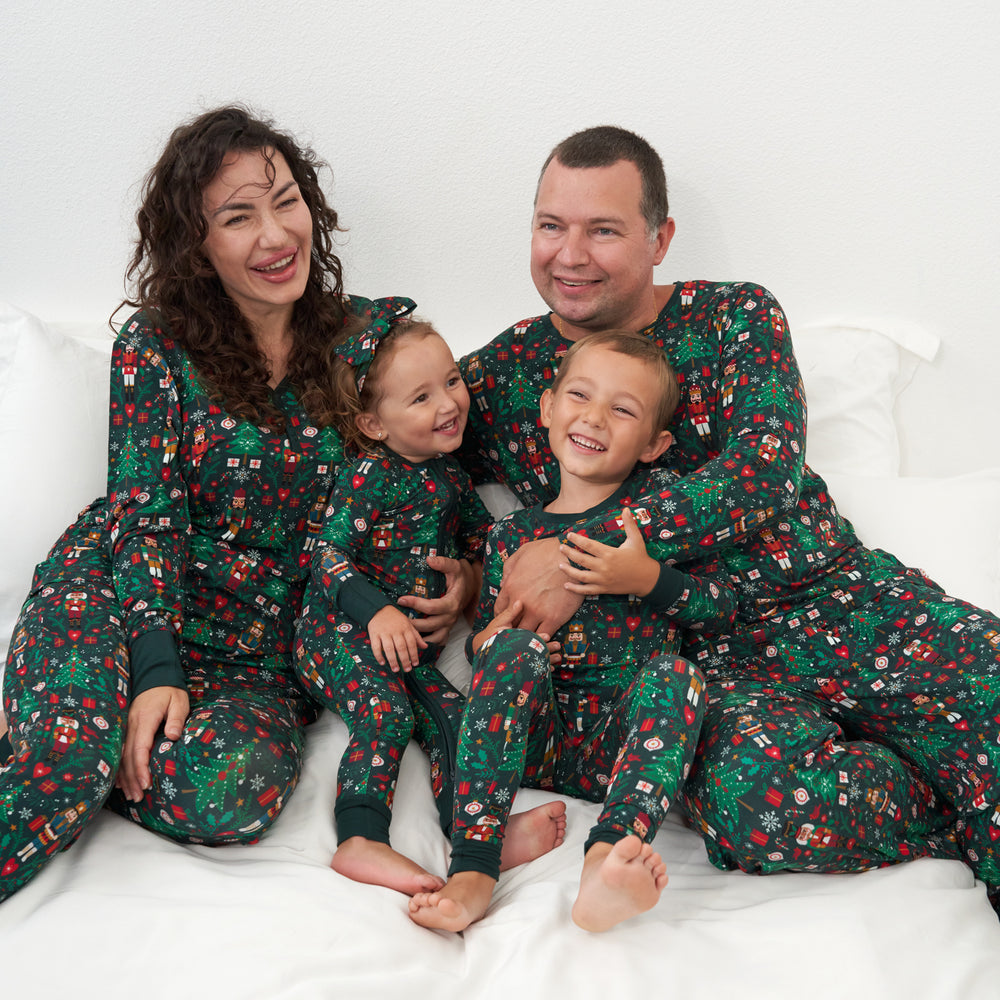 Family of four wearing matching Night at the Nutcracker Pajama sets. Mom is wearing women's Night at the Nutcracker women's pajama tops and matching women's pajama bottoms. Dad is wearing Night at the Nutcracker men's pajama top and matching pajama bottoms. Children are wearing a Night at the Nutcracker two piece pajama sets paired with a matching luxe bow headband.