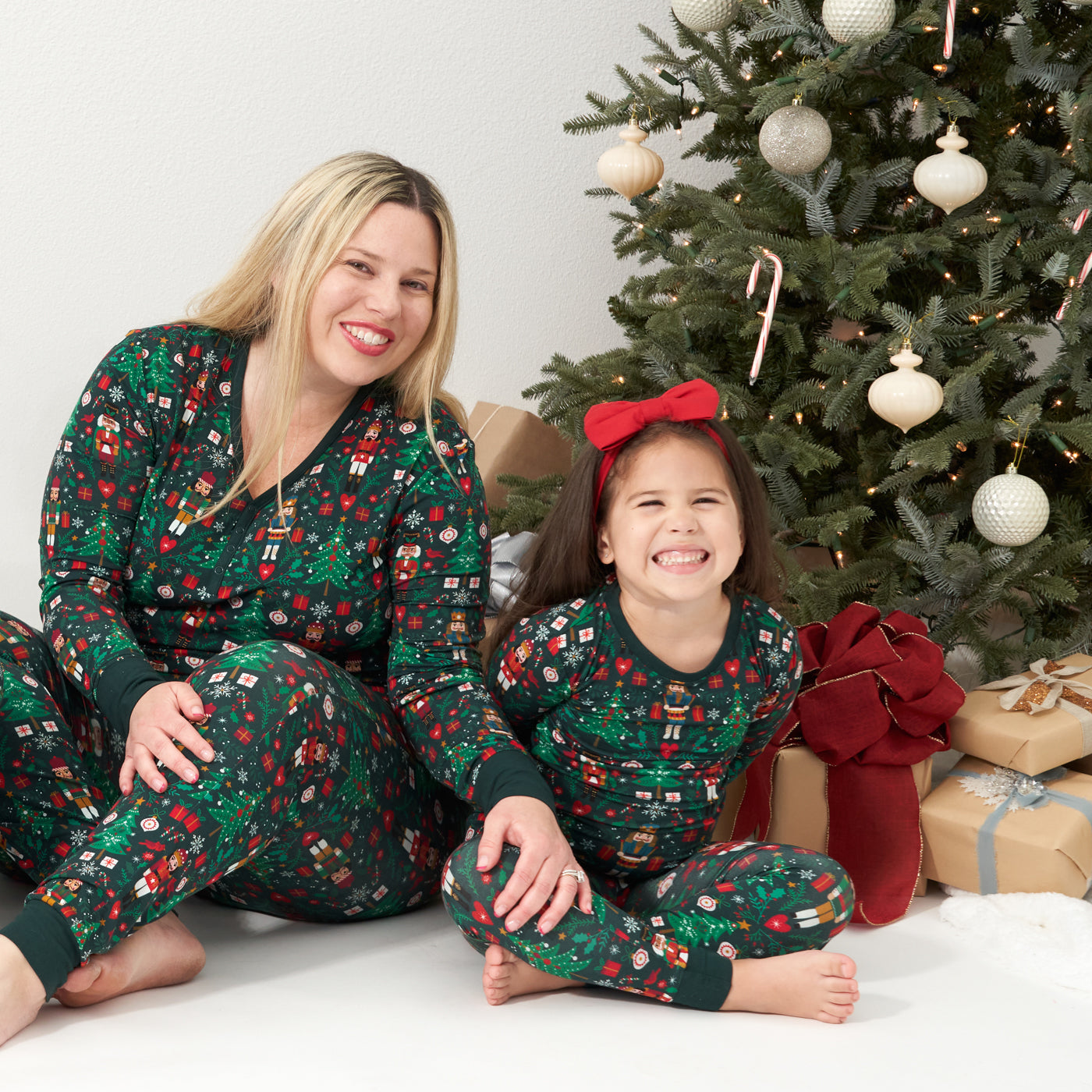 Mother and daughter posing wearing matching Night at the Nutcracker pajama sets. Mom is wearing a Night at the Nutcracker women's pajama top paired with women's pajama bottoms. Her child is wearing Night at the Nutcracker two piece pajama set paired with a Holiday Red luxe bow headband