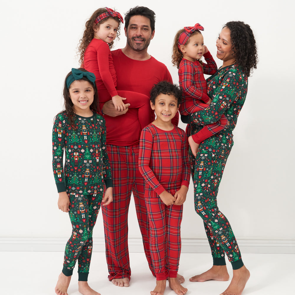 Mom is wearing Night at the Nutcracker women's pajama top and matching pajama bottoms. Dad is wearing Holiday Plaid men's pajama bottoms paired with a Holiday Red men's pajama top. Children are coordinating, one wearing Night at the Nutcracker two piece pajama set paired with an Emerald luxe bow headband. Two are wearing Holiday Plaid two piece pajama sets, paired with a Holiday Plaid luxe bow headband. One is wearing a Holiday Red two piece pajama set paired with a Holiday Plaid luxe bow headband. 