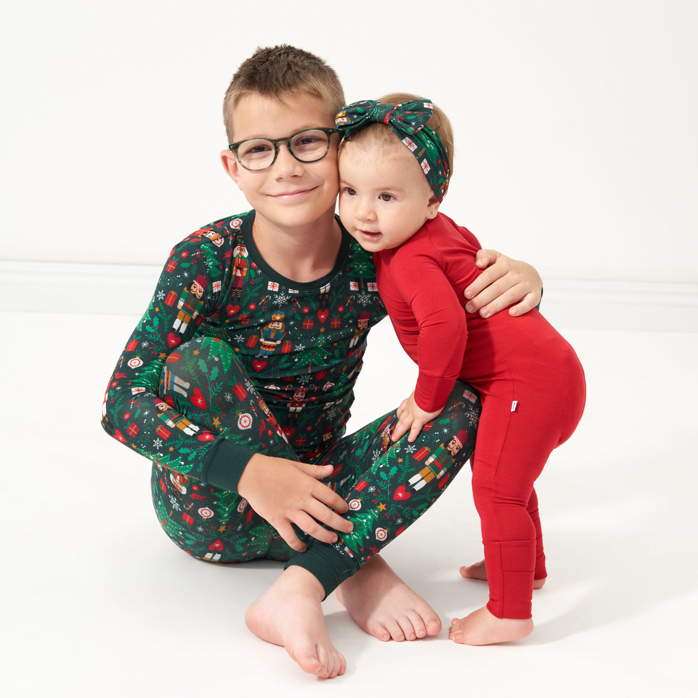 Two children sitting together. One child is wearing a Night at the Nutcracker two piece pajama set. Their sibling is wearing a Holiday Red zippy paired with a Night at the Nutcracker luxe bow headband