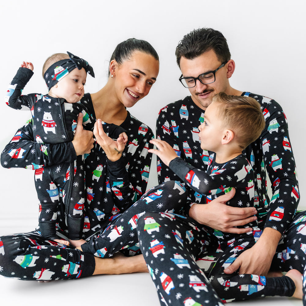 Family of four together wearing matching Polar Bear Pals pajama sets. Mom is wearing women's Polar Bear Pals pajama top paired with matching women's pajama pants. Dad is wearing men's Polar Bear Pals pajama top paired with matching men's pajama pants. And their children are wearing Polar Bear Pals pajamas in two piece and zippy styles paired with a matching luxe bow headband