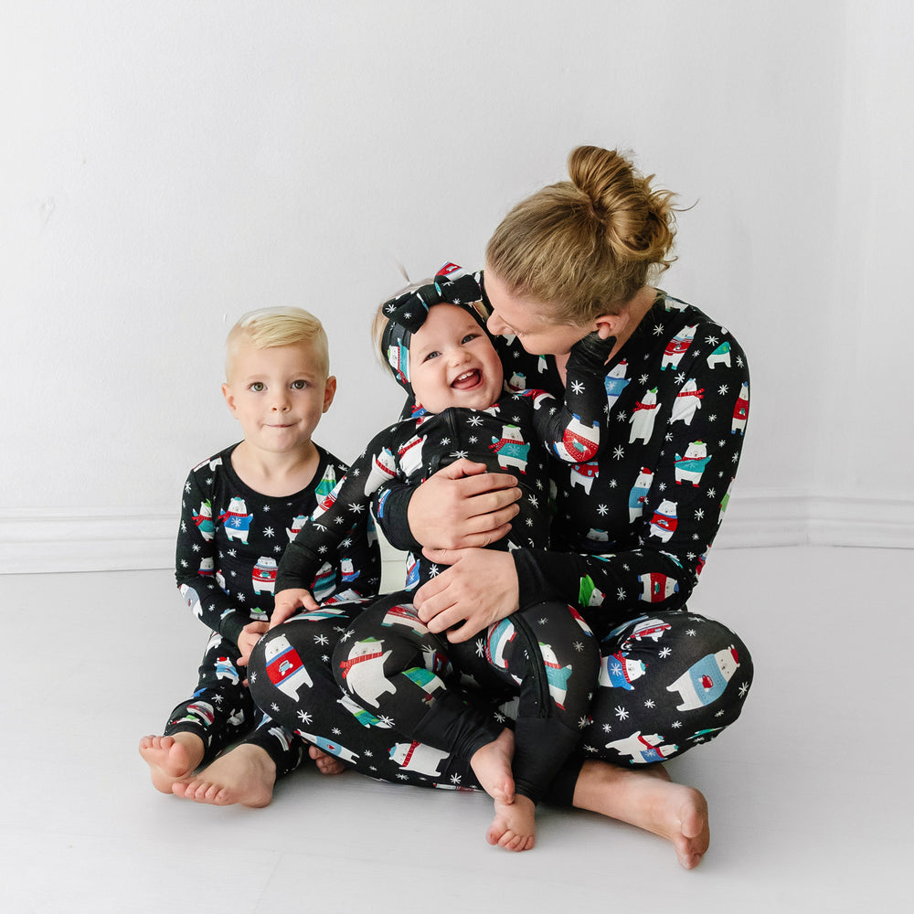 Mother and her two children wearing matching Polar Bear Pals pajamas. Mom is wearing Polar Bear Pals women's pajama top paired with matching women's pajama pants. Her children are wearing Polar Bear Pals printed pajamas in two piece pajama set and zippy styles paired with a matching luxe bow headband.