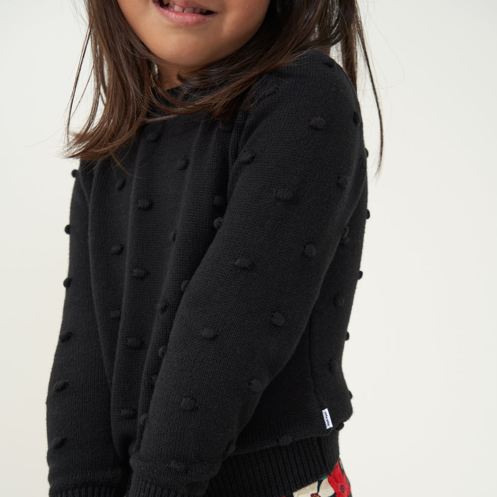 Close up image of a child wearing a Black pom pom sweater