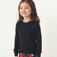 Close up image of a child looking to the side wearing a Black pom pom sweater