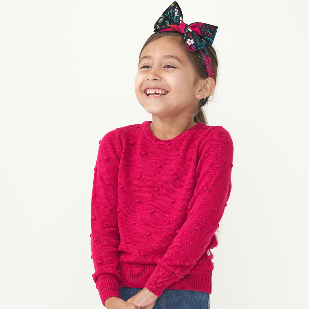 Child posing wearing a Mixed Berry Pom Pom sweater paired with a Berry Merry luxe bow headband