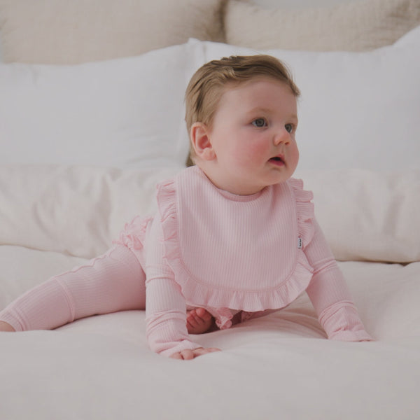 Baby sitting on a bed wearing the Prima Pink Ribbed Ruffle Bib
