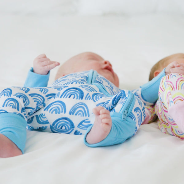 Two children on a bed wearing coordinating Blue Rainbows and Pastel Rainbows zippies