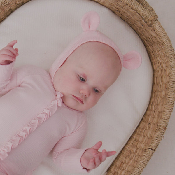 Baby in a basket crib wearing the Prima Pink Bear Bonnet