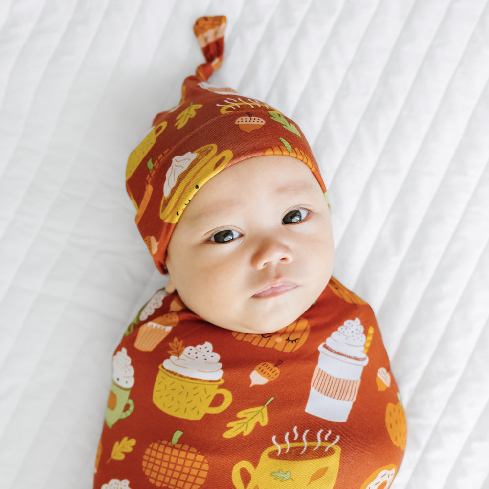 Close up image of a child laying on a bed swaddled in a Pumpkin Spice swaddle and hat set