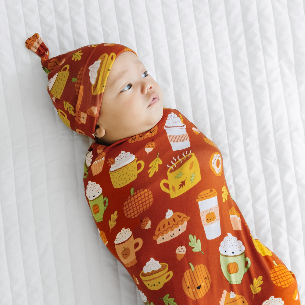 Side view image of a child laying on a bed swaddled in a Pumpkin Spice swaddle and hat set