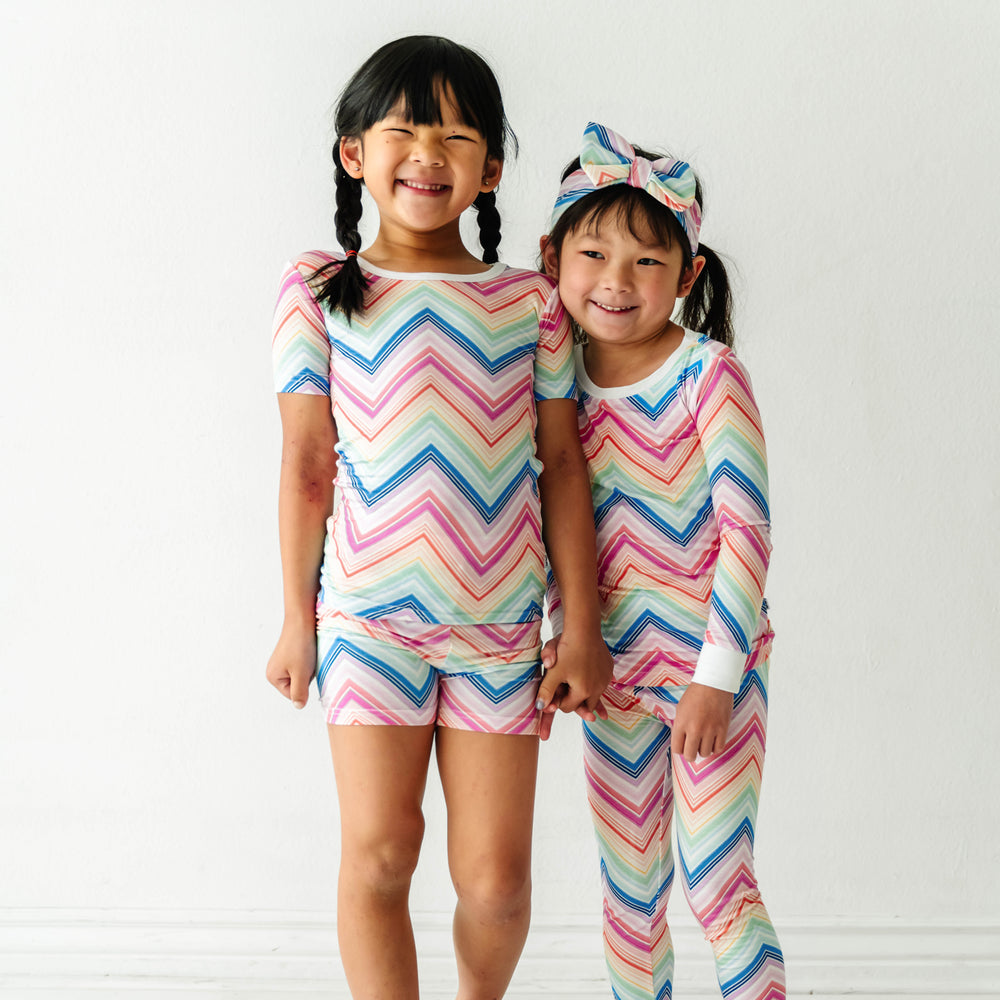 Click to see full screen - Two children holding hands wearing matching Rainbow Chevron printed short sleeves and shorts pajama set and long sleeve pajama set