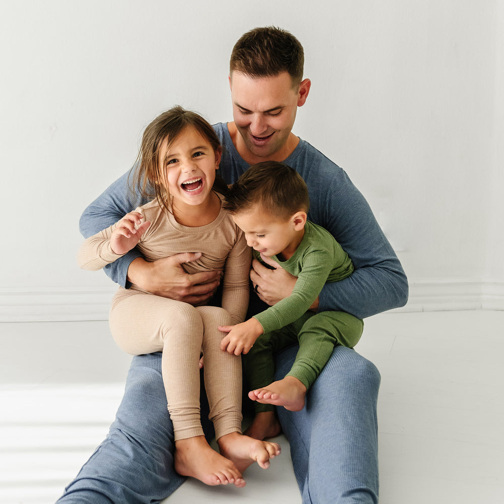 Father and his two children. Dad is wearing Heather Dusty Indigo Ribbed men's pajama top and pants. His children are wearing two piece pajamas in Heather Cypress Ribbed and Heather Fawn Ribbed colors