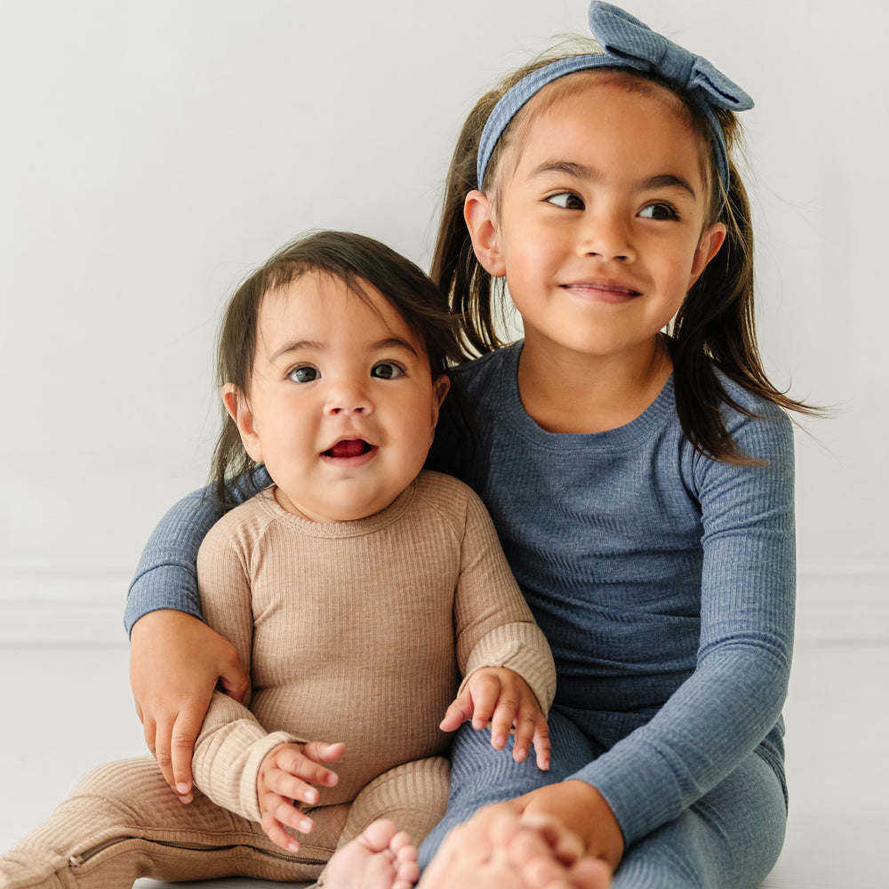 Two children sitting with their arms linked wearing coordinating Heather Fawn Ribbed crescent zippy and a Heather Dusty Indigo two piece pajama set paired with a matching luxe bow headband