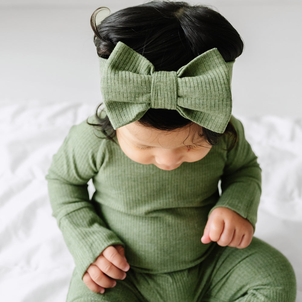 Close up image of a child wearing a Heather Cypress Green Ribbed luxe bow headband and matching crescent zippy