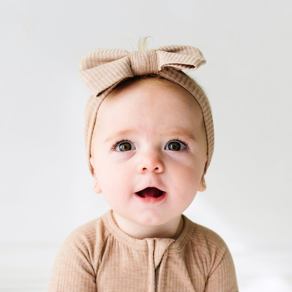 Close up image of a child wearing a Heather Fawn ribbed luxe bow headband paired with a matching zippy