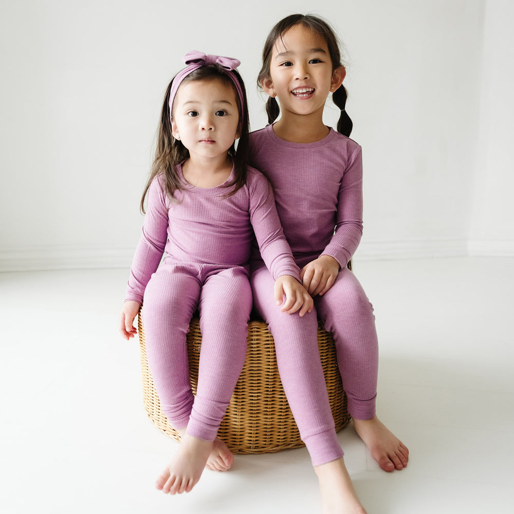 Two children sitting wearing Heather Mulberry ribbed two piece pajama sets paired with a matching luxe bow headband