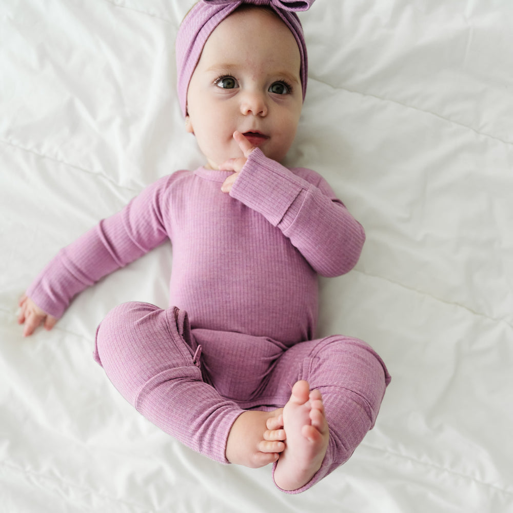 Child laying on a blanket wearing a Heather Mulberry Ribbed Crescent Zippy paired with a matching luxe bow headband