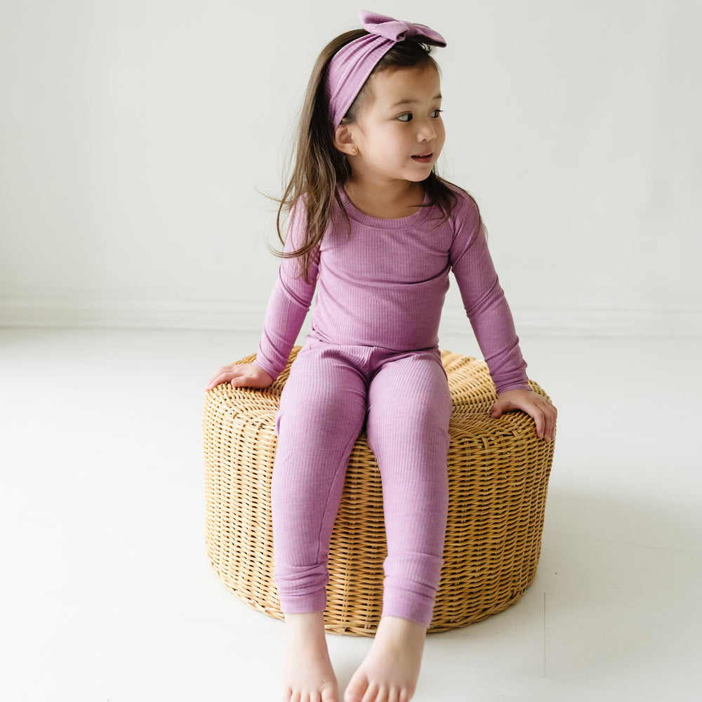 Child sitting wearing a Heather Mulberry ribbed two piece pajama set paired with a matching luxe bow headband