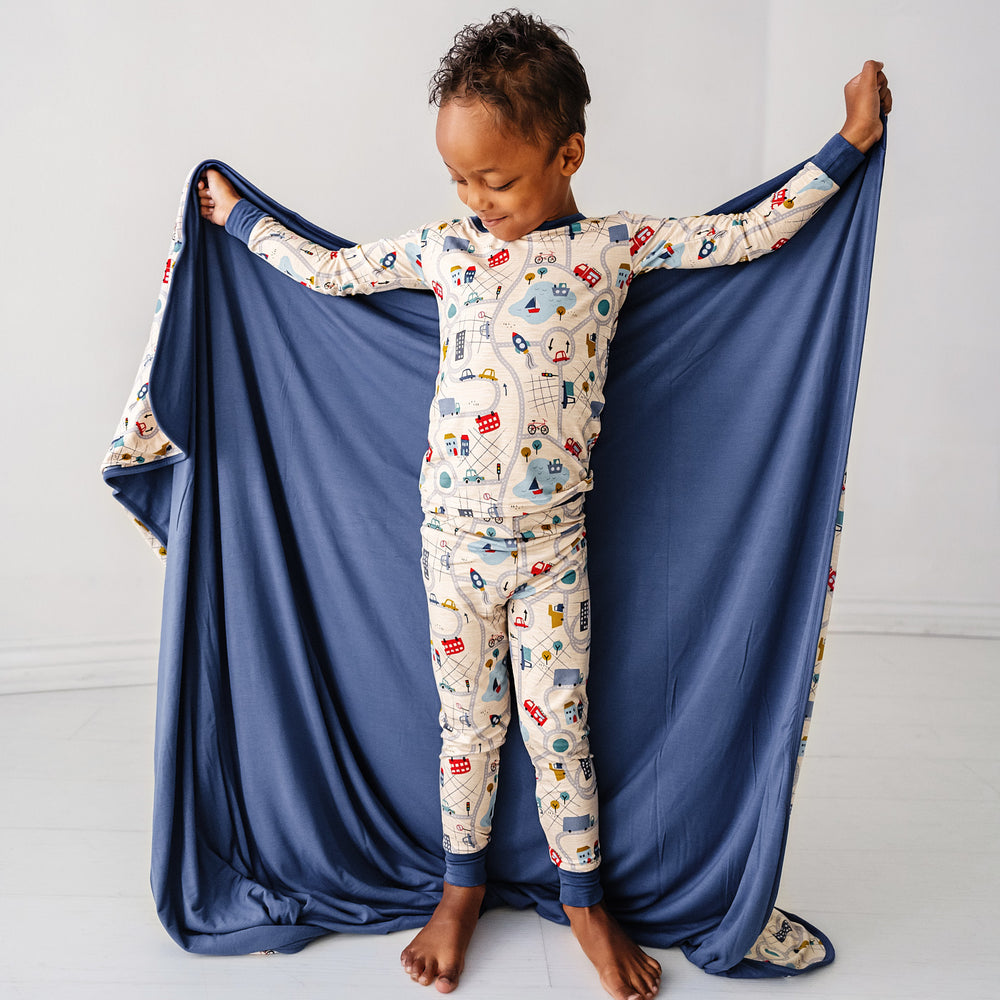 Click to see full screen - Child wearing a Blue Road Trip two piece pajama set holding up a matching large cloud blanket showing the solid blue backing