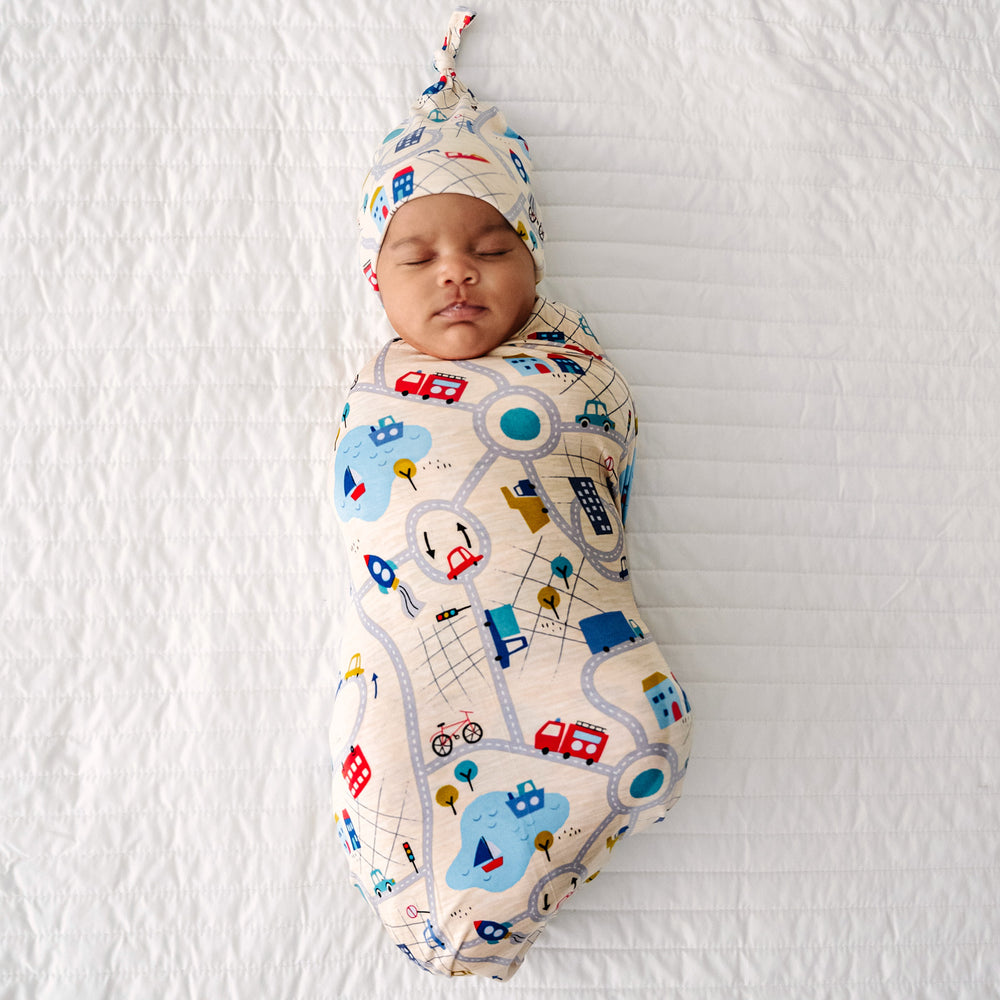 Click to see full screen - Child laying on a bed swaddled in a Blue Road Trip swaddle and hat set