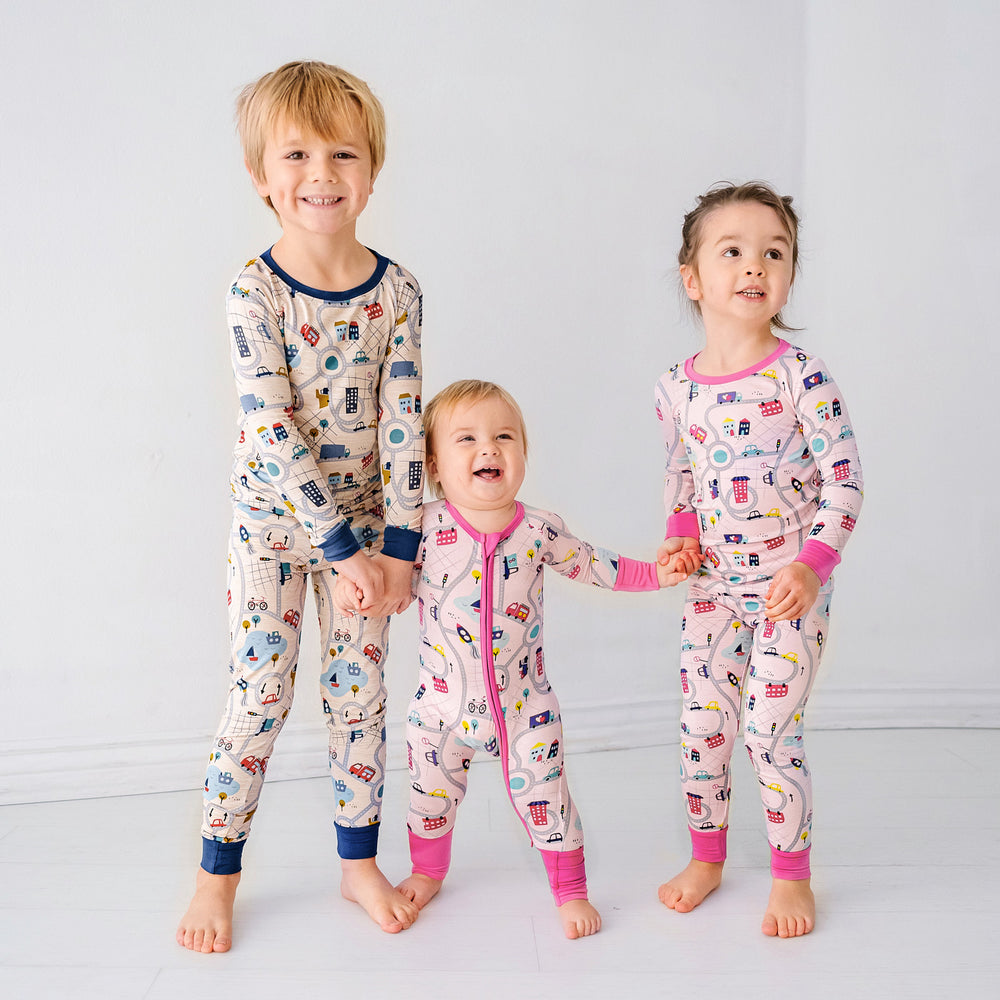 Click to see full screen - Three children holding hands wearing coordinating Road Trip pajama sets. Two children are wearing Blue and Rosy Road Trip two piece pajama sets and one child is wearing a Rosy Road Trip zippy