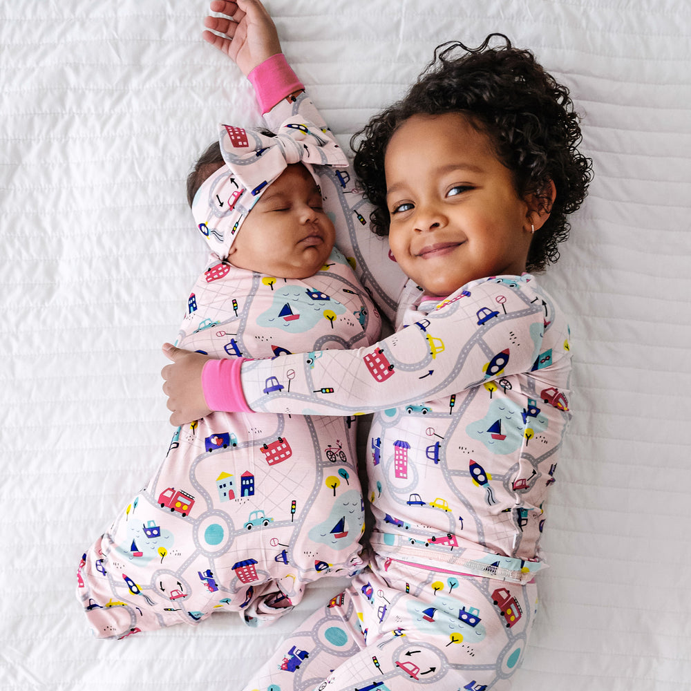 Click to see full screen - Two children laying together. One child is laying on a bed swaddled in a Rosy Road Trip swaddle and luxe bow headband set and her sister is wearing a matching Rosy Road Trip two piece pajama set