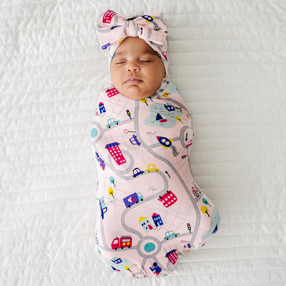 Click to see full screen - child laying on a bed swaddled in a Rosy Road Trip swaddle and luxe bow headband set