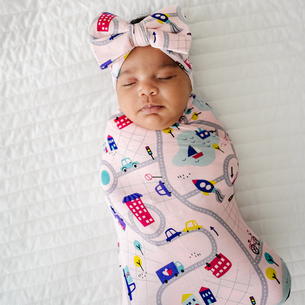 Click to see full screen - Close up image of a child laying on a bed swaddled in a Rosy Road Trip swaddle and luxe bow headband set