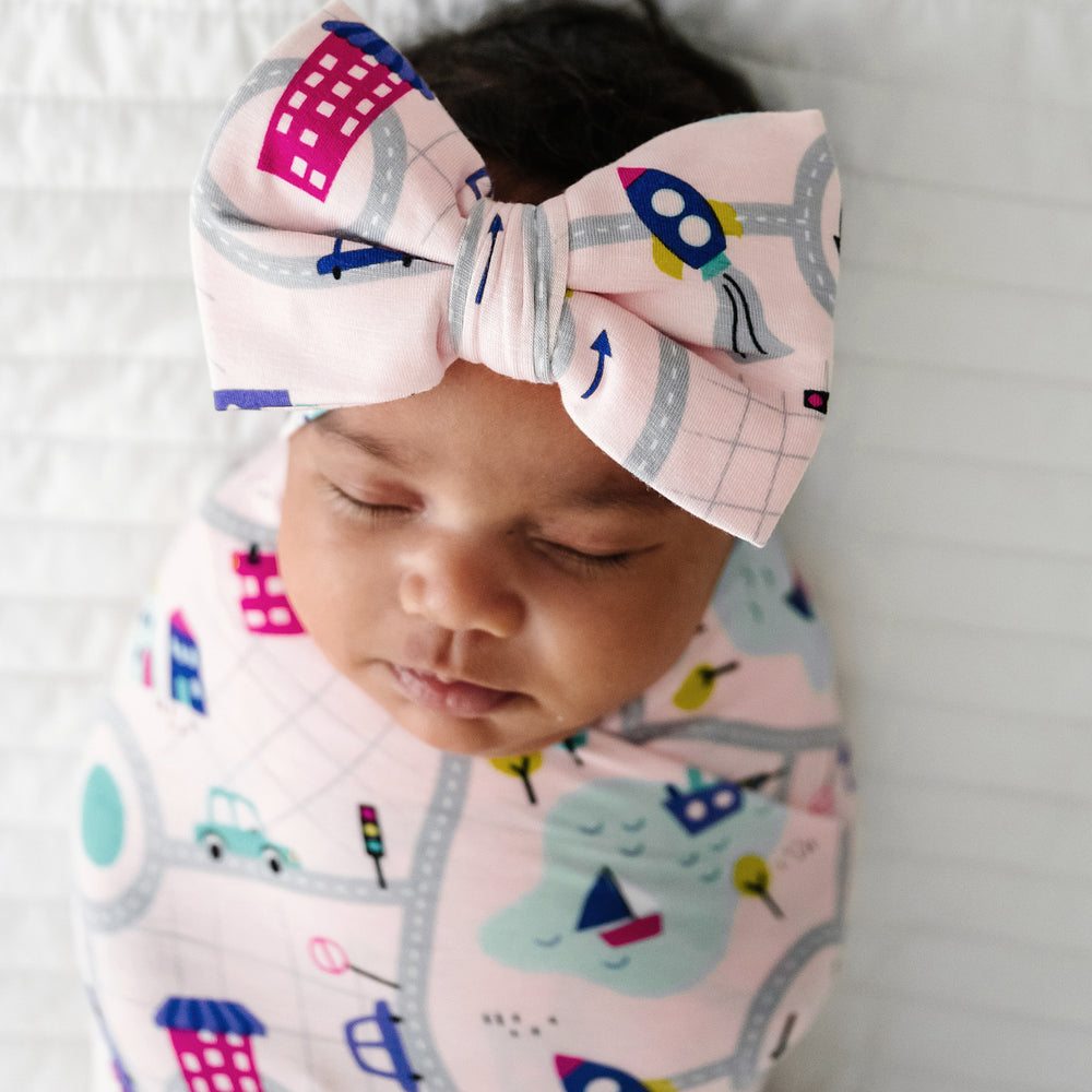 Click to see full screen - Close up image of a child laying on a bed swaddled in a Rosy Road Trip swaddle and luxe bow headband set focusing on the luxe bow headband