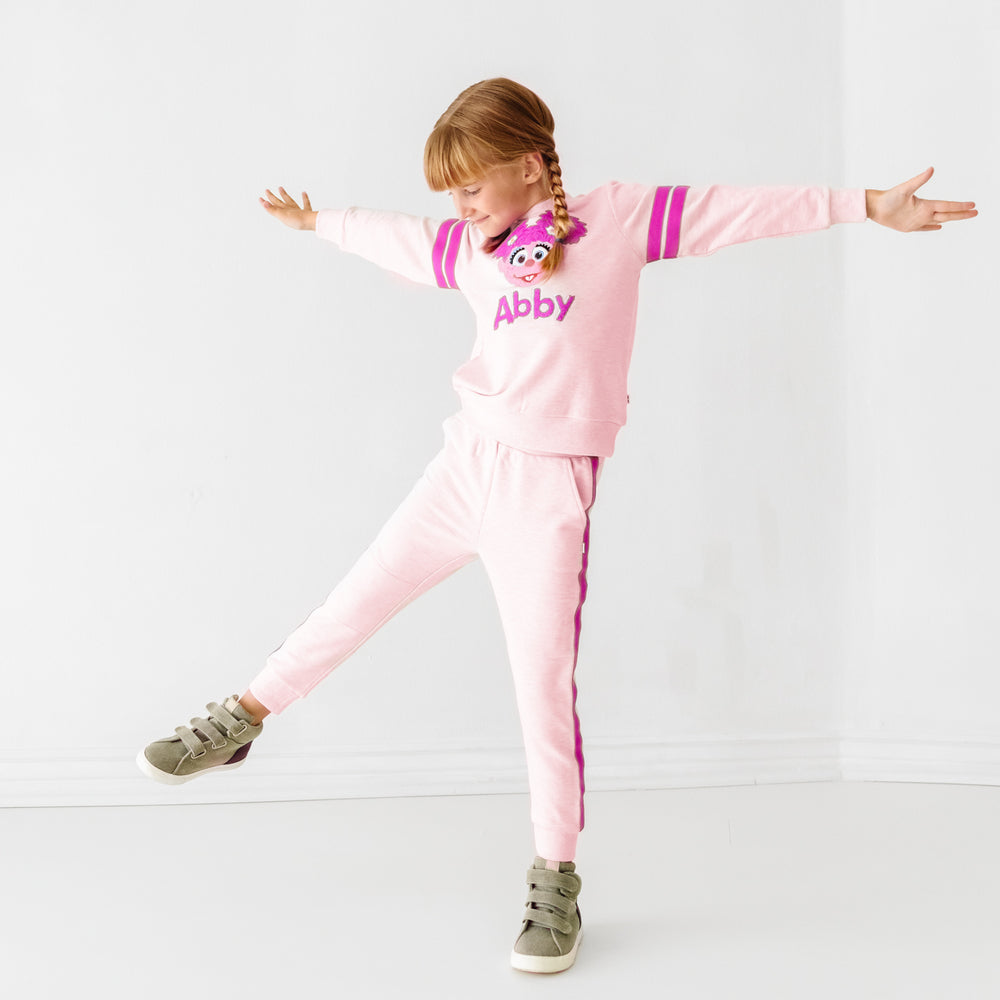 Child standing on one foot wearing a Sesame Street Abby crewneck sweatshirt and jogger set