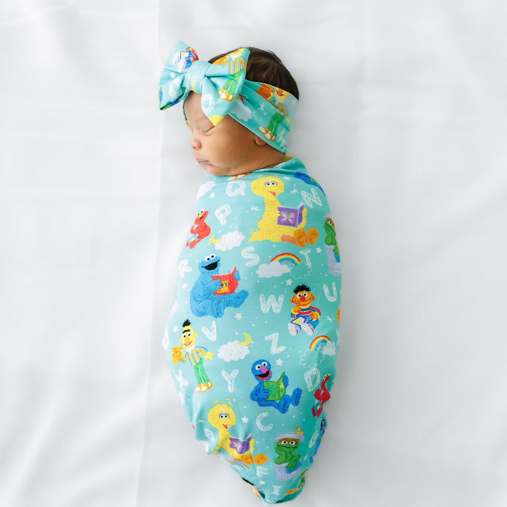Child laying on a bed swaddled in a Spelling with Sesame Street Swaddle and Luxe Bow Headband Set