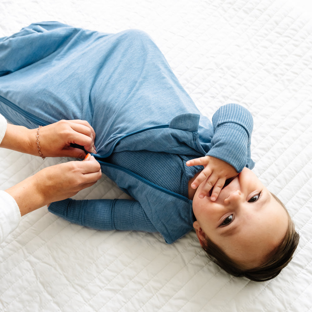 Click to see full screen - Child laying on a blanket wearing a Heather Blue Sleepy Bag being zipped up by a parent