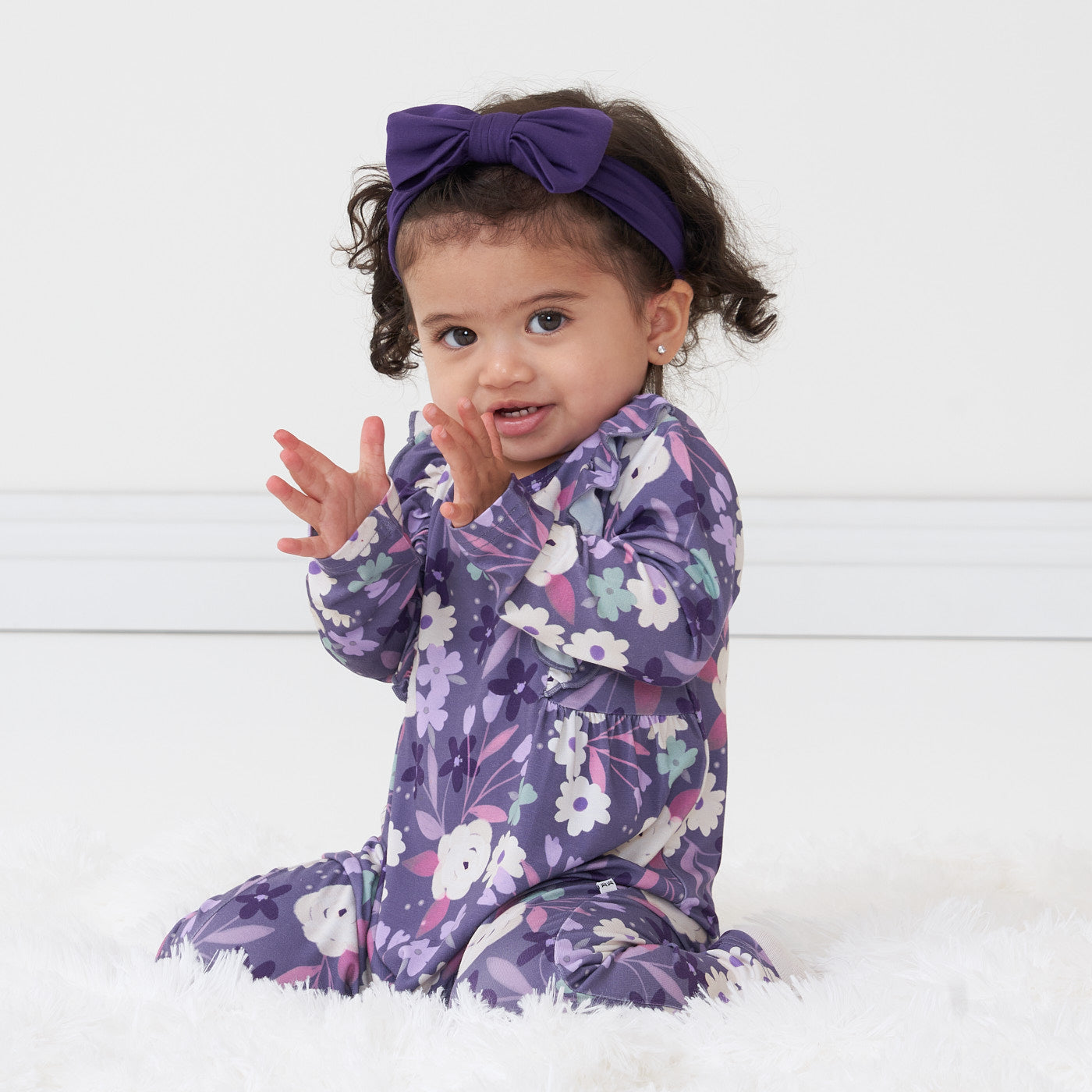 Child sitting wearing a Sugar Plum Floral ruffle bib romper paired with a Deep Amethyst luxe bow headband