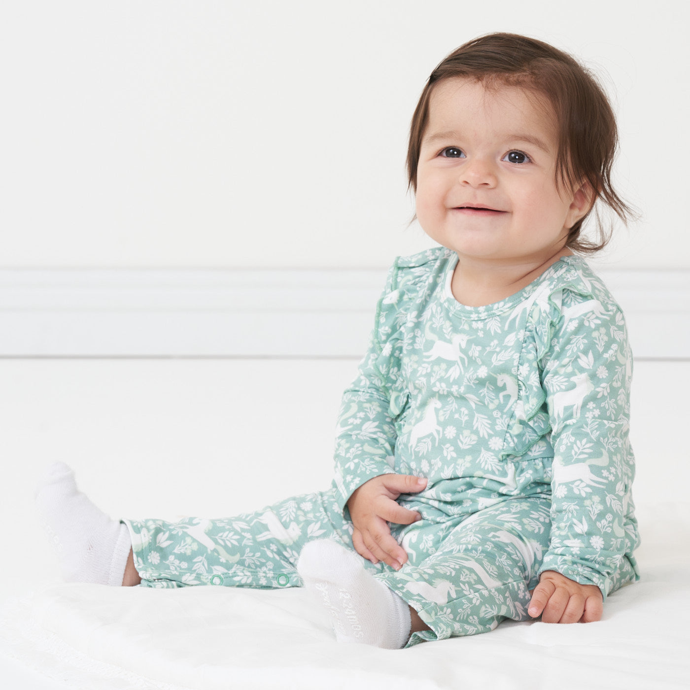 Side view image of a child sitting on the ground wearing a Unicorn Garden ruffle bib romper