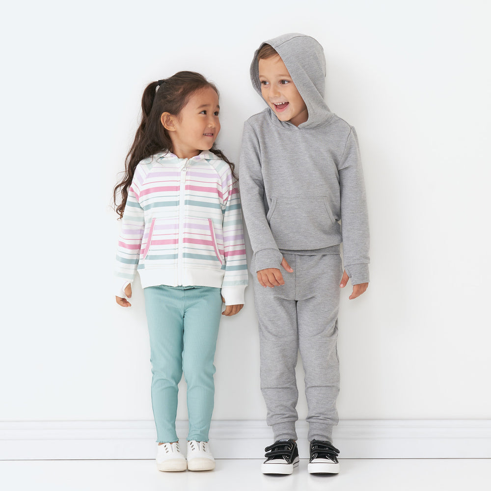 Two children posing together. wearing winter play collection styles. One child is wearing Aqua Mist Ribbed cozy lettuce leggings paired with a Winter Stripe zip hoodie and the other is wearing a Heather Gray pull over hoodie paired with matching joggers