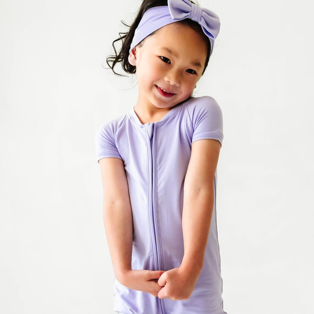 Child posing wearing a Wisteria shorty romper paired with a matching Wisteria luxe bow headband