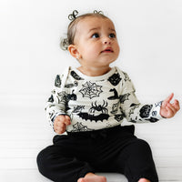 Child sitting wearing a Witches Brew Bodysuit paired with Black joggers