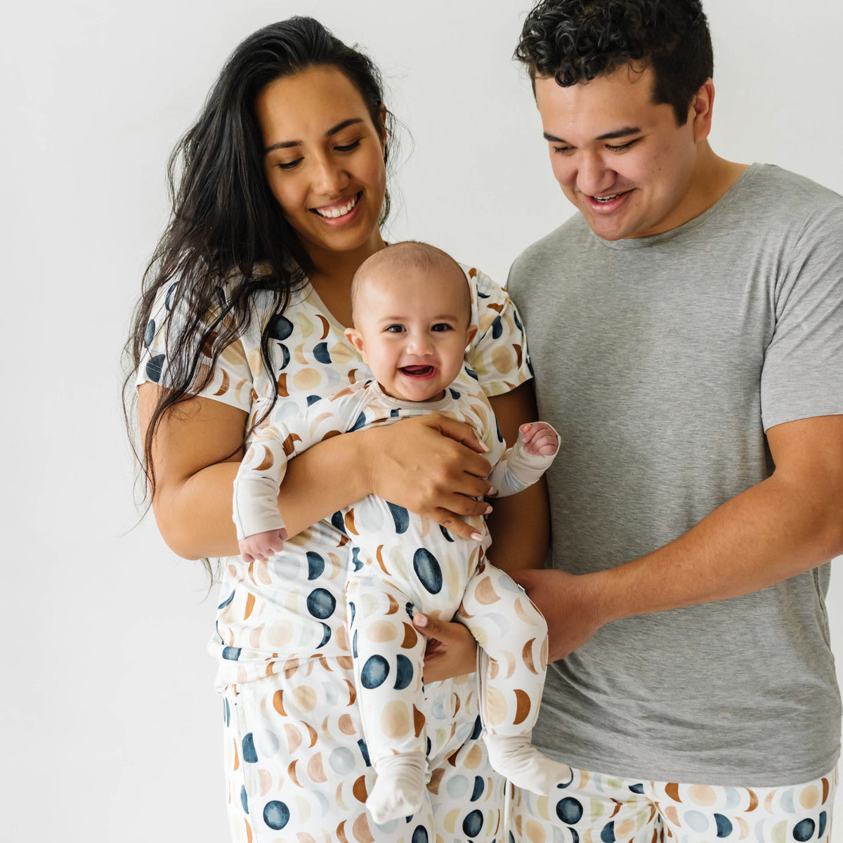 family of three posing together coordinating Luna Neutral pajamas. Mom is wearing a Luna  Neutral women's short sleeve pj top paired with matching women's shorts. Dad is wearing a Heather Gray men's short sleeve pj top paired with Luna Neutral pj shorts. Child is wearing a matching crescent zippy