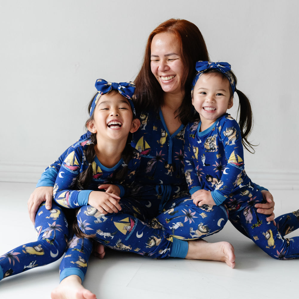 Mother and her two children wearing matching pajamas. Mom is wearing women's Where the Wild Things Are women's pajama top and matching pants. Children are matching Where the Wild Things Are two piece pajama sets and matching luxe bow headbands