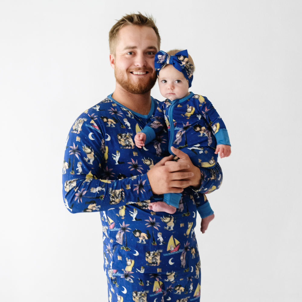 Close up image of a man wearing Where the Wild Things Are men's pajama top holding his daughter wearing a matching zippy and luxe bow headband