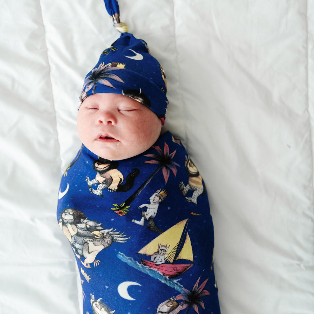 Close up image of a child laying on a bed swaddled in a Where the Wild Things Are swaddle and hat set