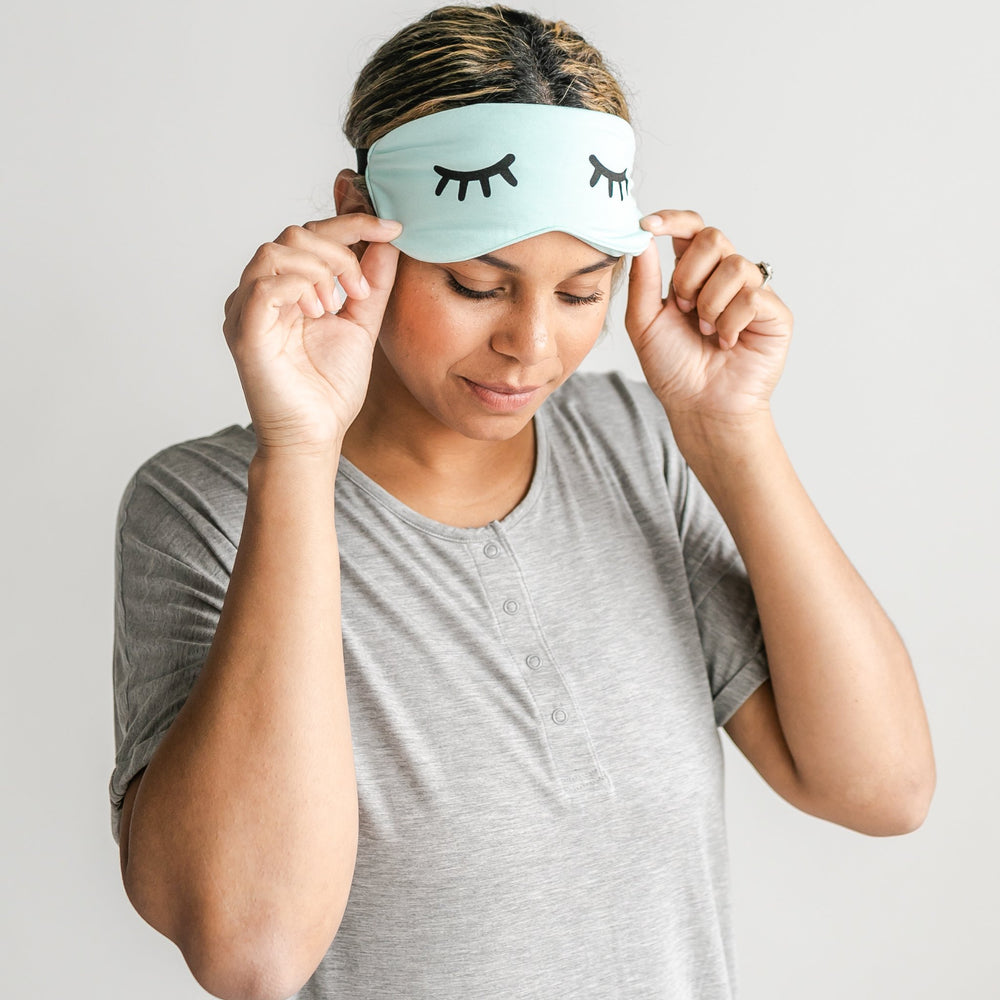 Image of female model wearing an adult sleeping eye mask in aquamarine. The eye mask features Little Sleepies eyes printed on it. 
