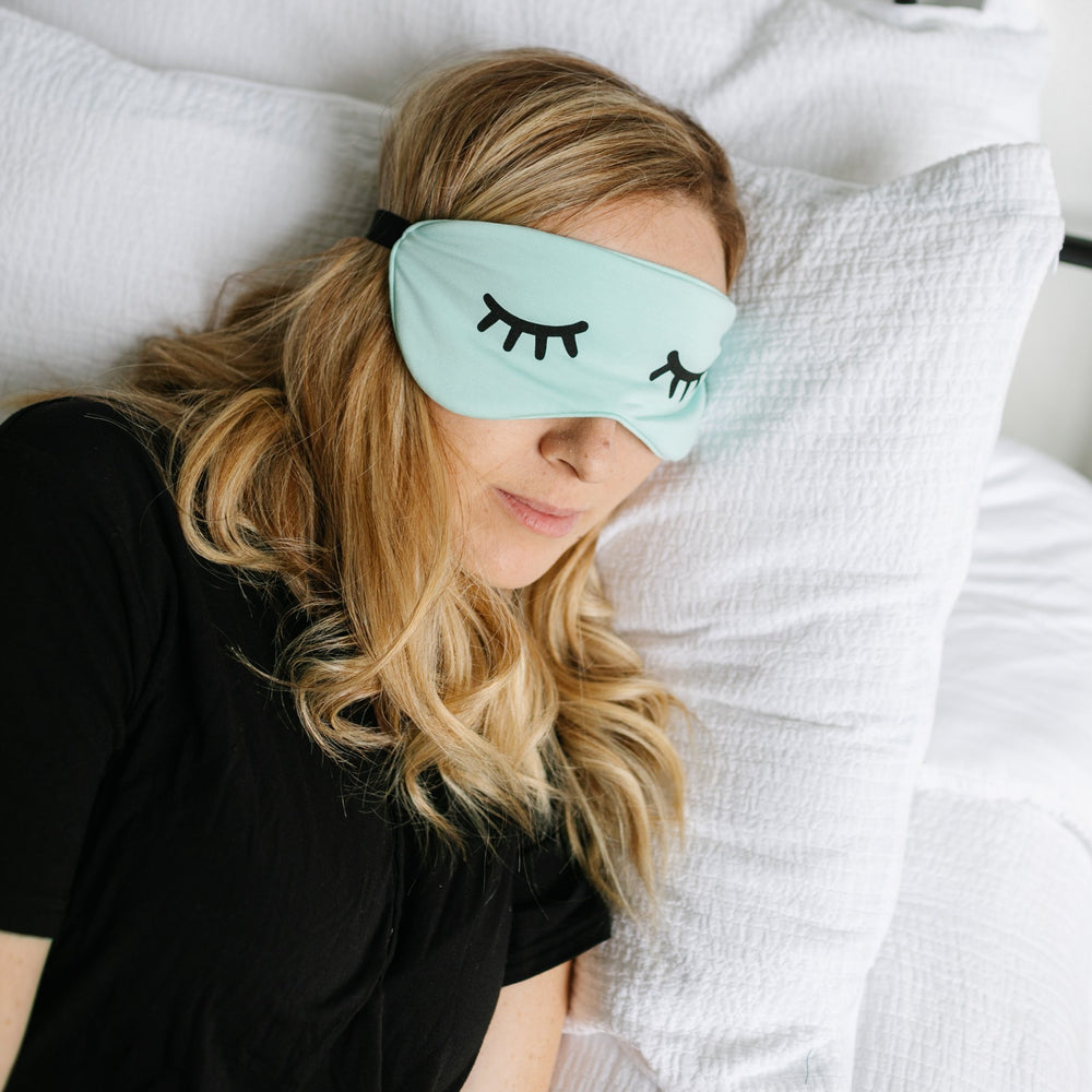 Image of female model wearing an adult sleeping eye mask in aquamarine. The eye mask features Little Sleepies eyes printed on it.