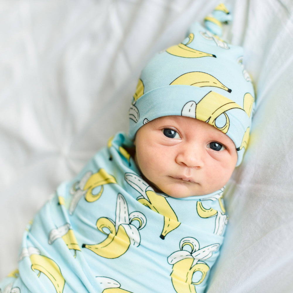 Baby boy wrapped up in banana printed swaddle. He is also wearing the matching banana printed hat. This set features a light blue background with pops of yellow coming from the bananas.