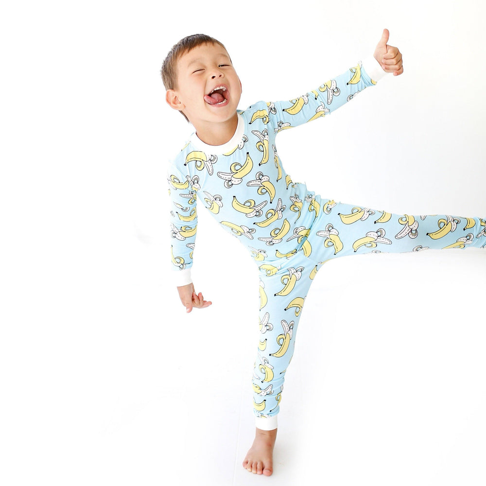 Little boy wearing banana printed pajama set. This print has a light blue background and white trim accents, with pops of yellow coming from the bananas. 