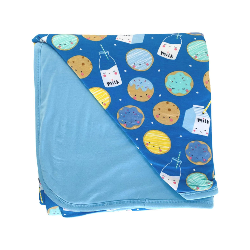 Flat lay image of triple layer large cloud blanket in Blue Cookies and Milk print. This blanket features a double-sided design, with one side that shows  a variety of different cookies with bottles and boxes of milk on a blue background and the other side featuring a coordinating solid light blue