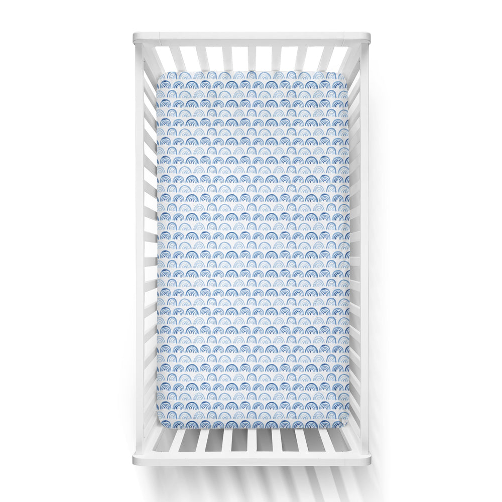Aerial view of a standard fitted infant crib sheet in Blue Rainbows print on a white crib